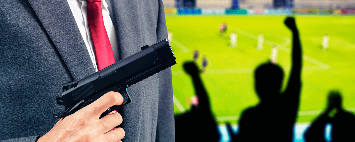Homem trajado de terno com arma na mão na arquibanca de um estádio de futebol.
